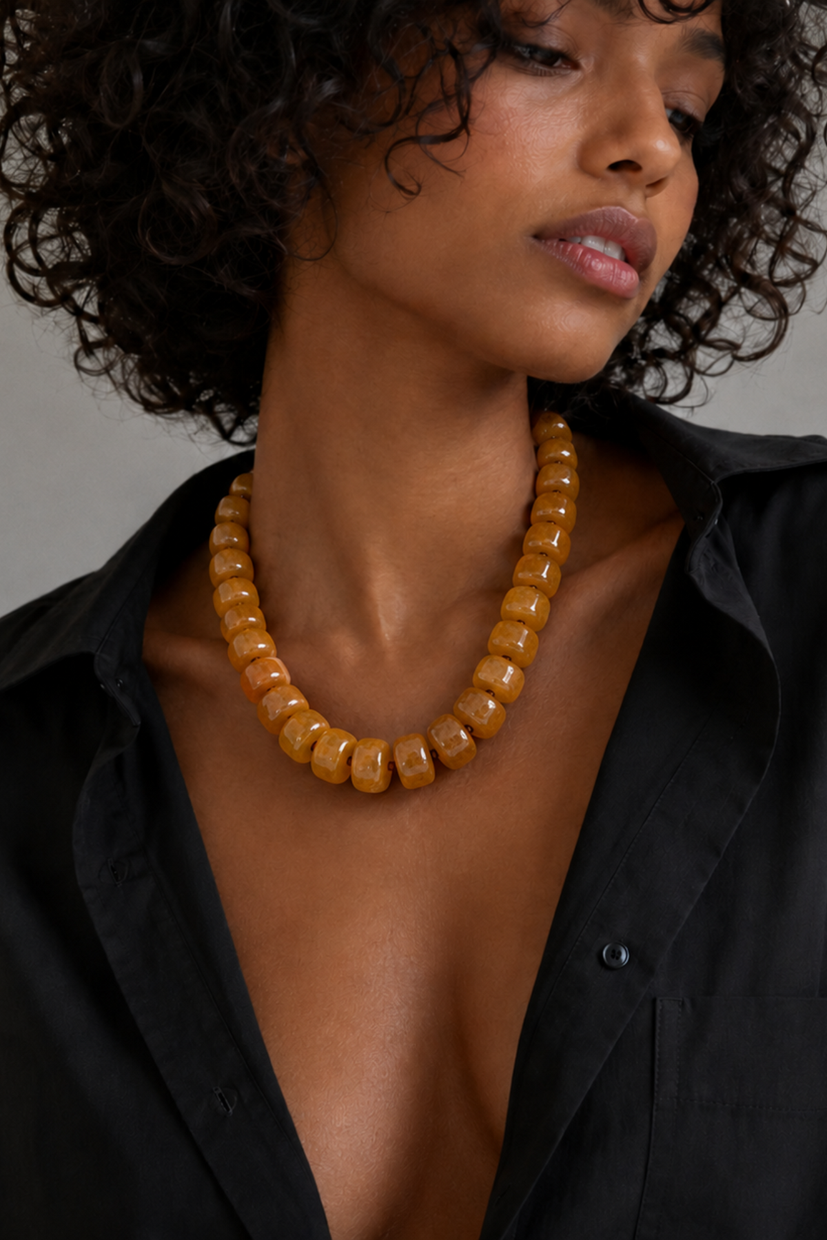 A close-up of a model wearing a chunky amber-brown beaded necklace with a black button-down shirt against a neutral gray background.