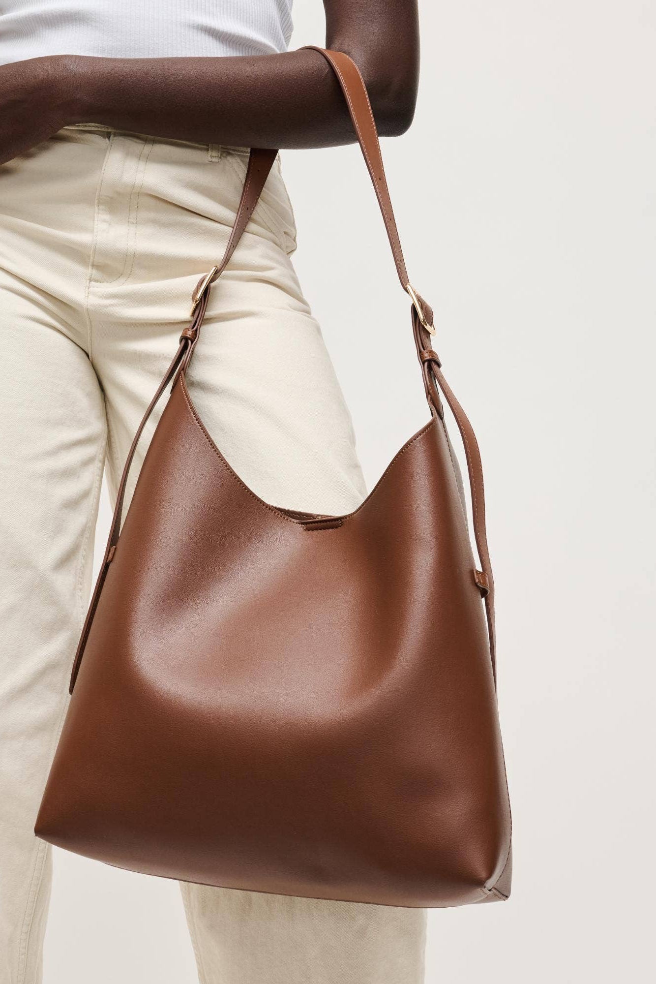 Brown leather handbag held by a person wearing beige pants on a light background