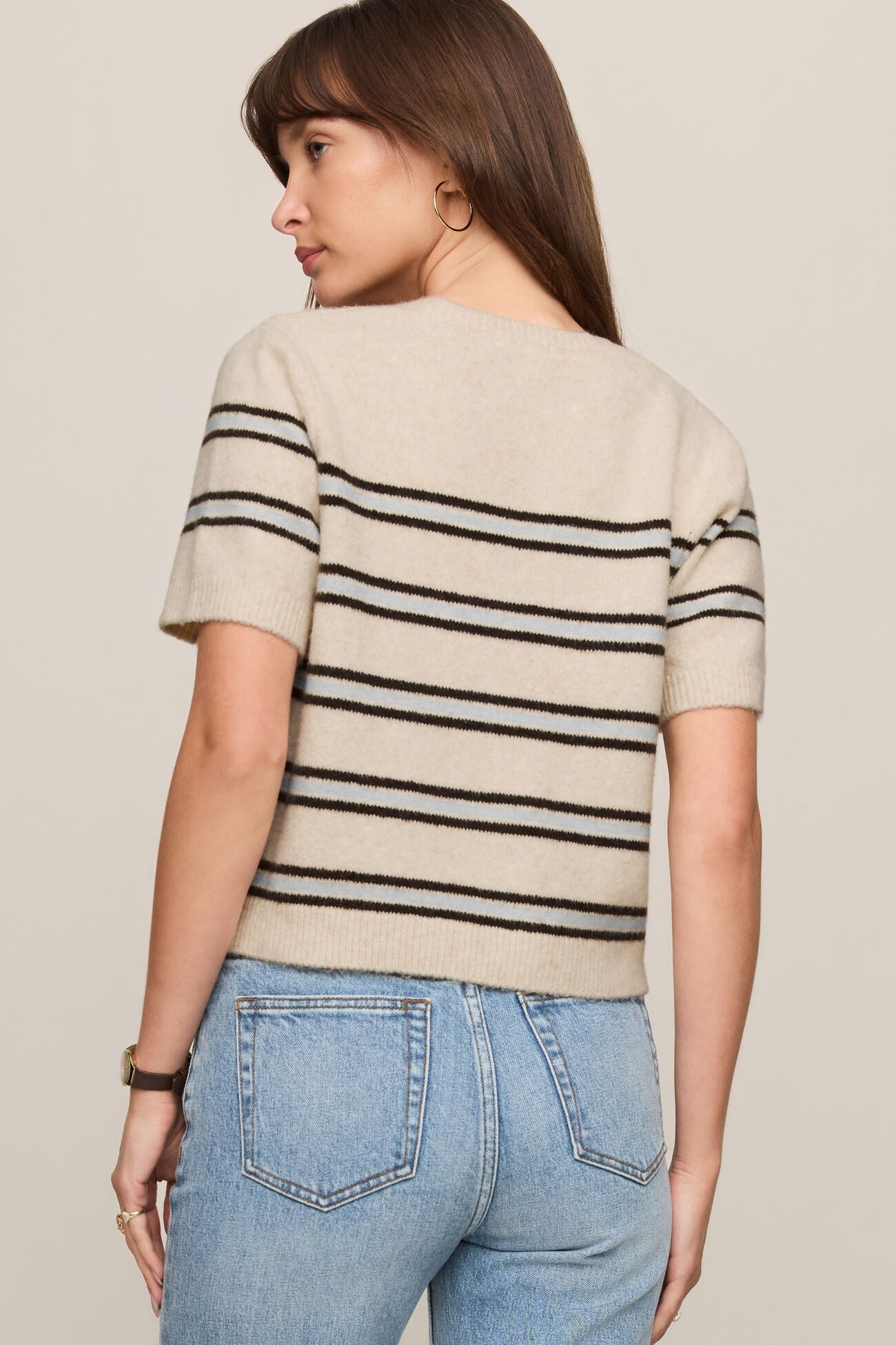 Woman wearing a beige sweater with black stripes and blue jeans on a plain background