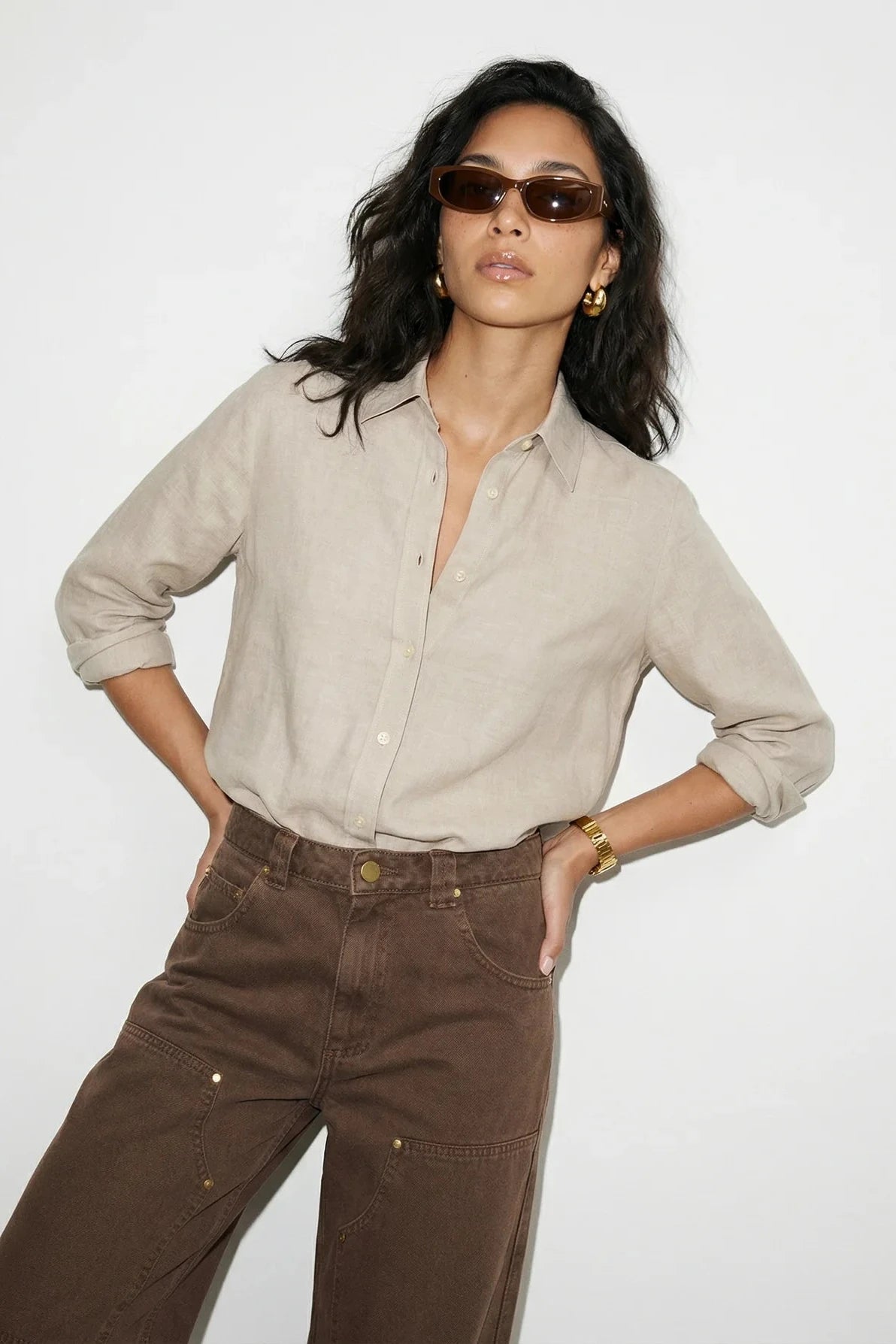 Woman wearing a beige shirt and brown pants with sunglasses on a white background
