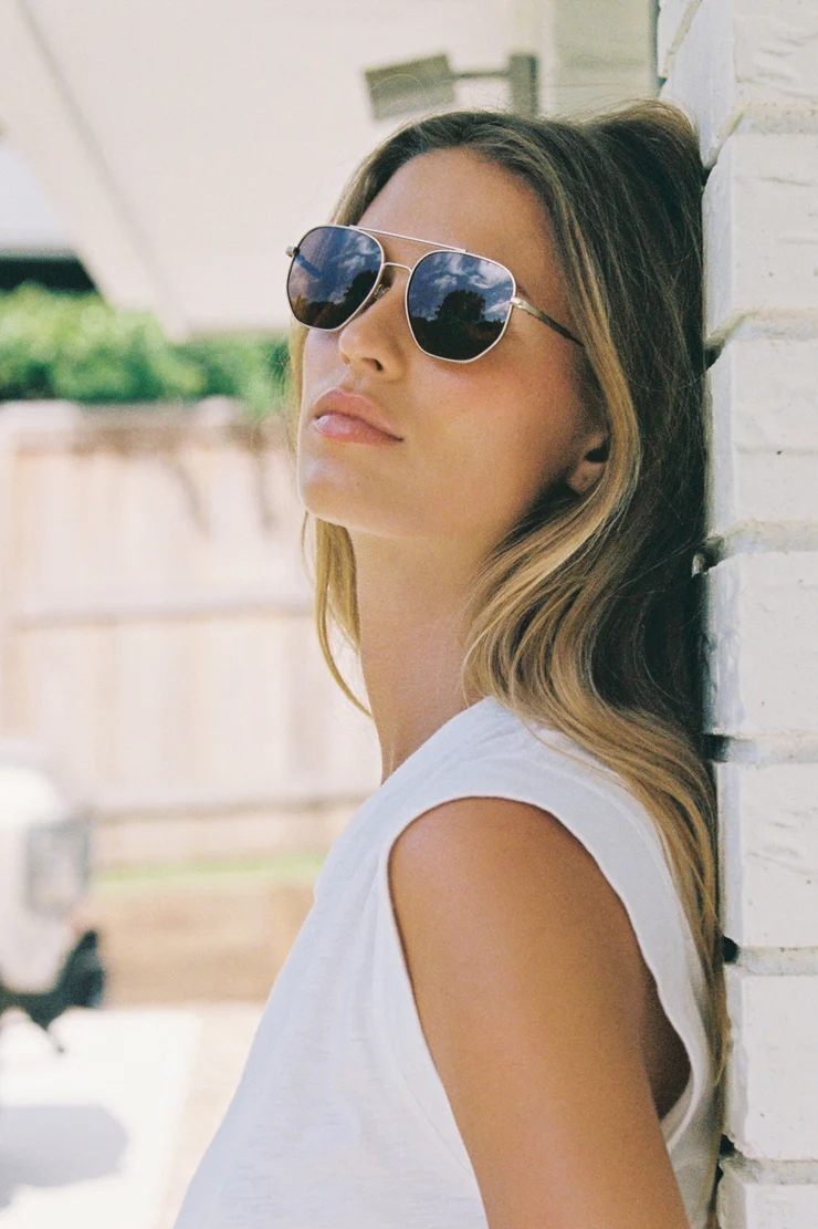 Woman wearing sunglasses leaning against a white wall outdoors