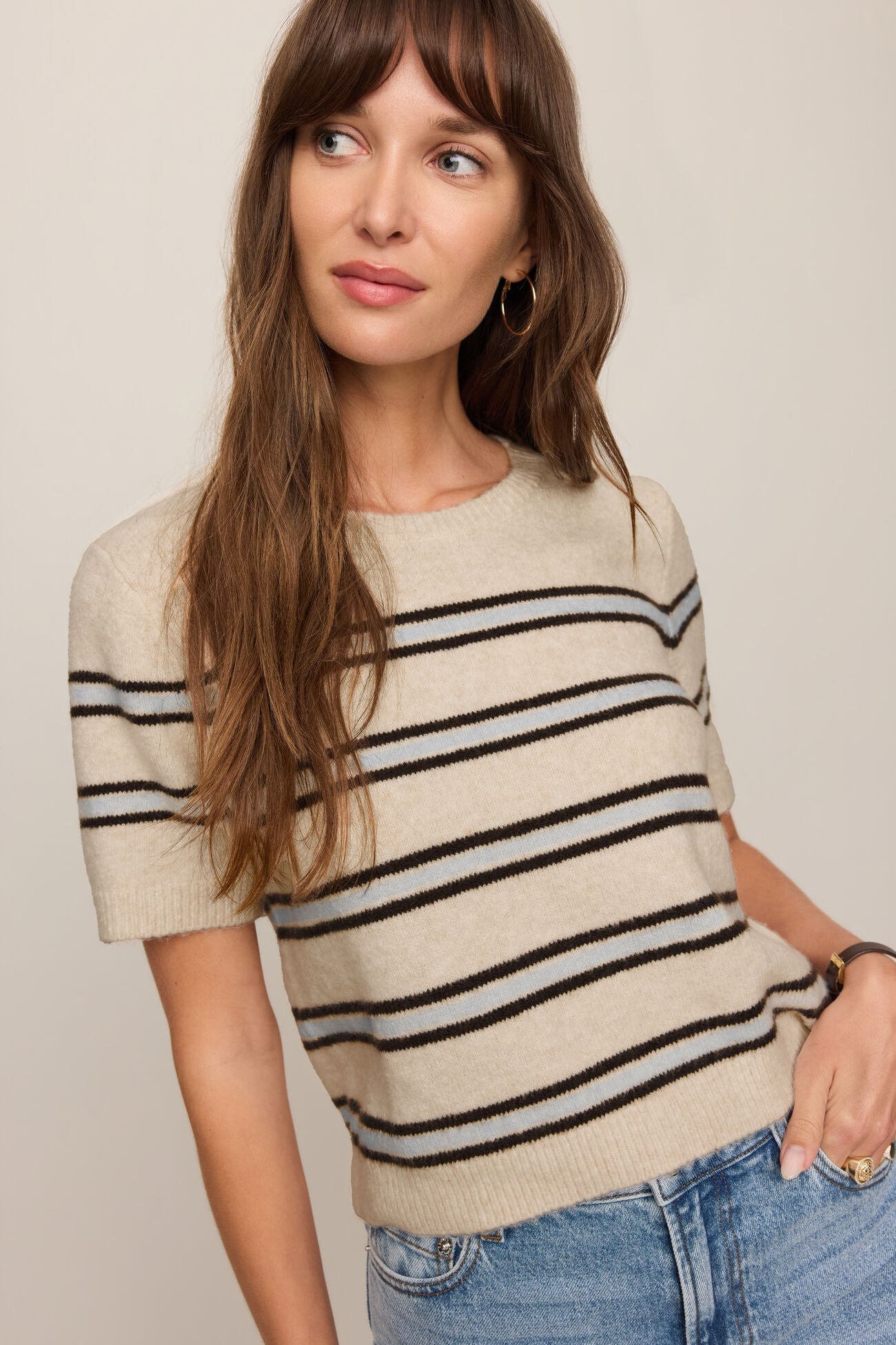 Woman wearing a beige and black striped sweater with blue jeans against a plain background