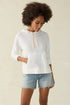 A female model wearing a white slub knit hoodie with a drawcord at the hood and a kangaroo pocket, paired with blue denim shorts.