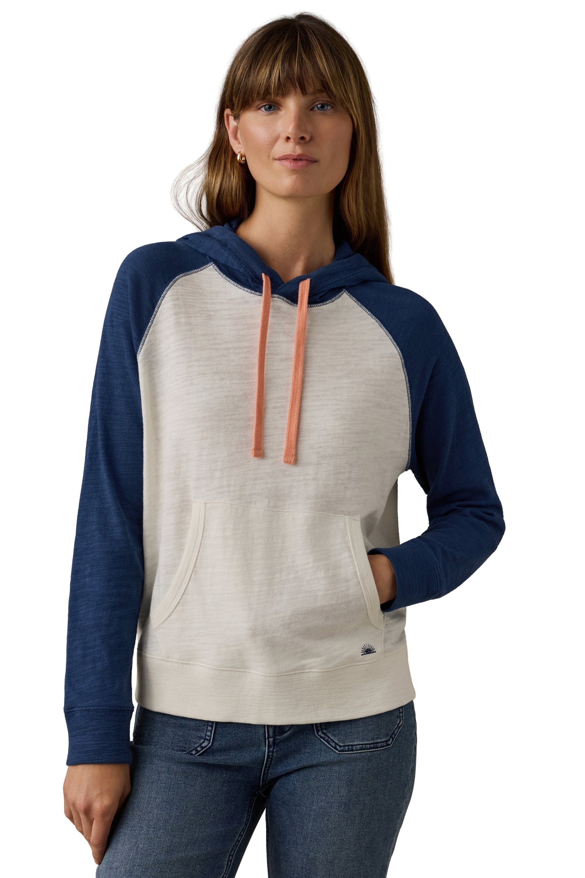 A woman wearing a two-tone raglan hoodie with a cream-colored body and navy blue sleeves and hood, featuring peach drawstrings, paired with blue jeans against a white background.