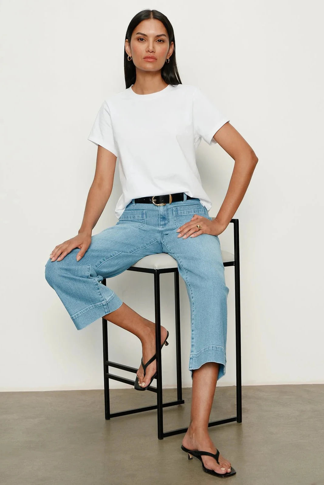 Woman sitting on a stool wearing a white t-shirt and light blue jeans against a plain background
