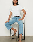 Woman sitting on a stool wearing a white t-shirt and light blue jeans against a plain background