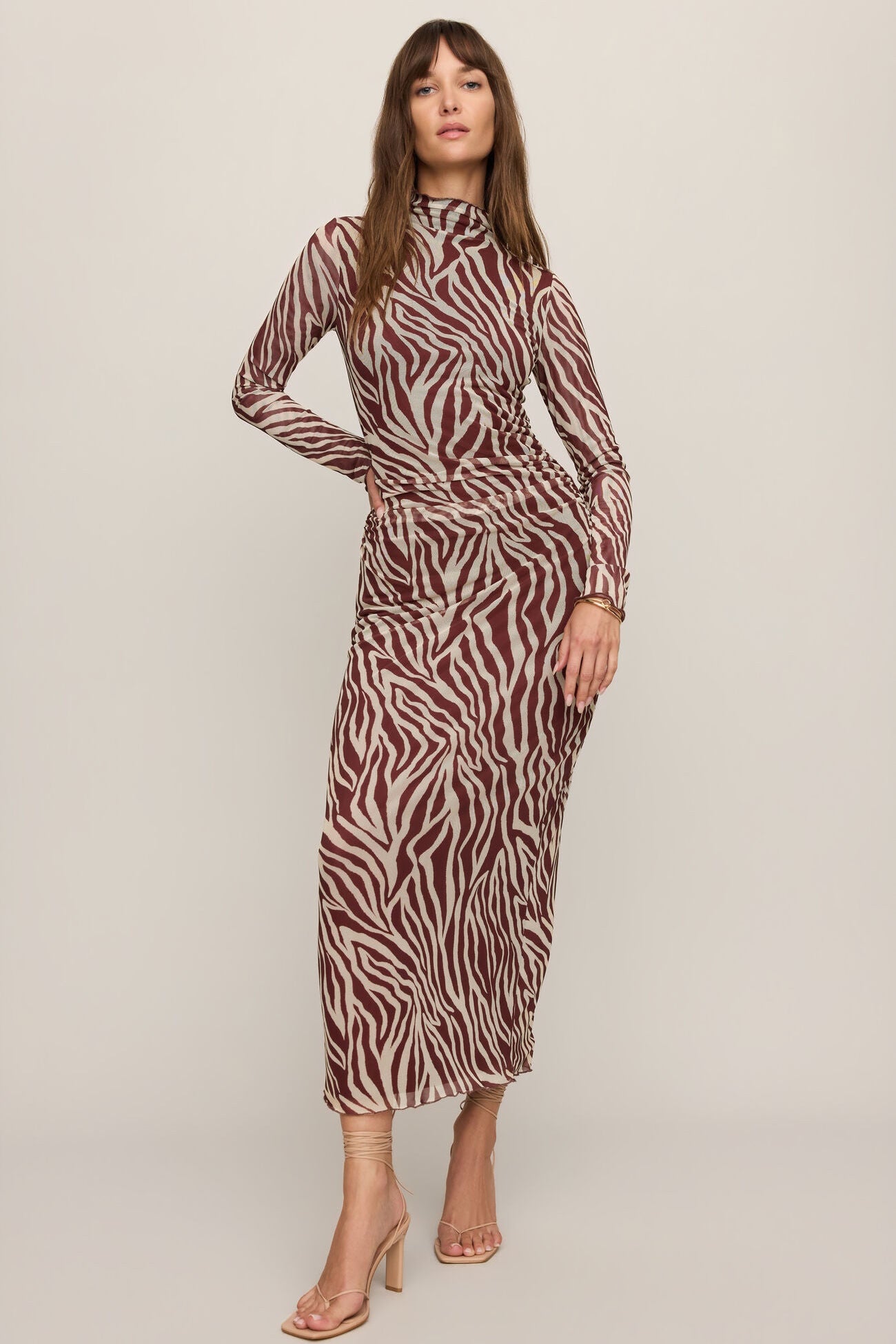 A woman modeling a long-sleeve high-neck midi dress with a brown and beige zebra print, paired with clear high-heeled sandals, against a solid light beige studio background.