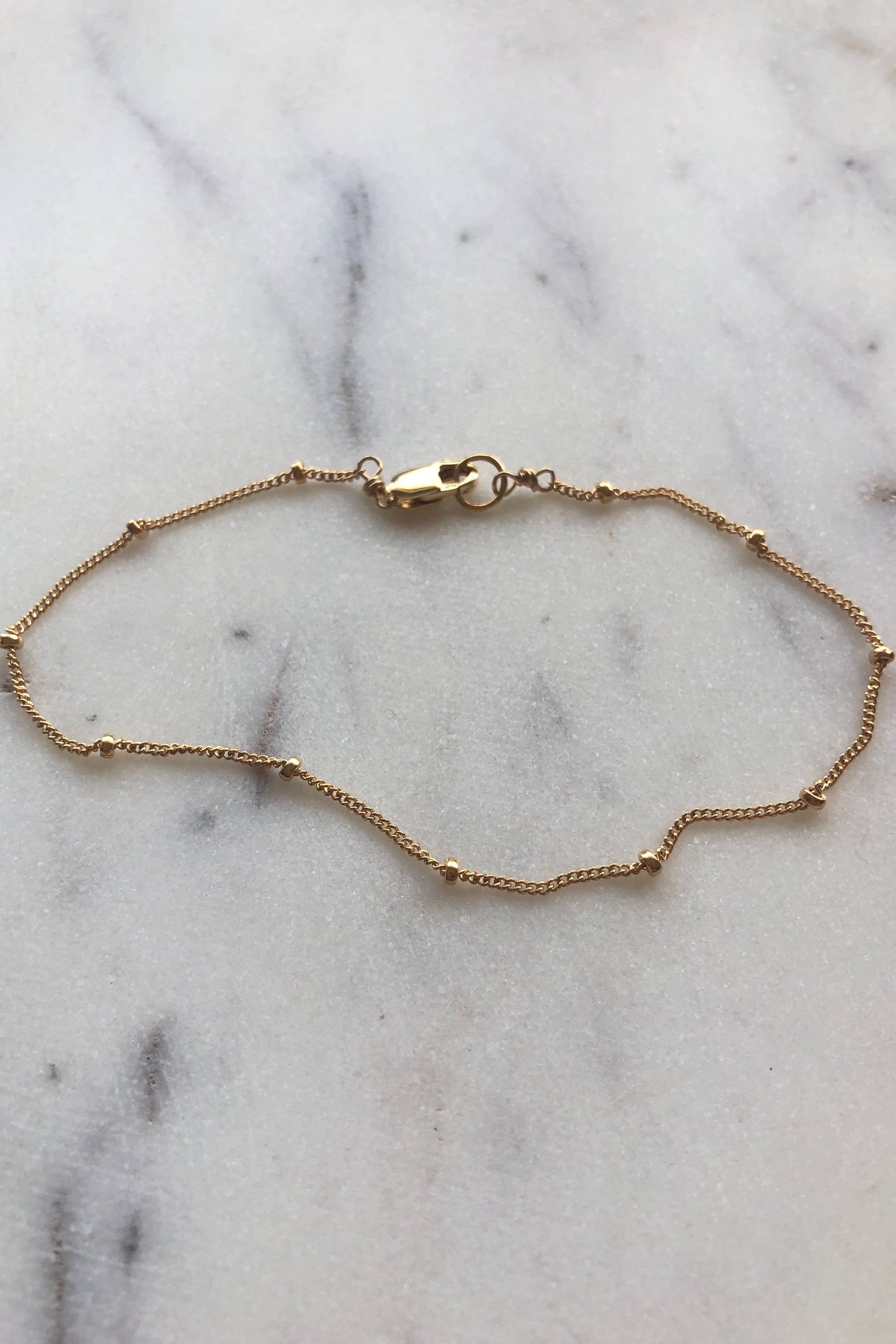 A delicate gold satellite chain bracelet with a total circumference of 7 inches, made of 14k gold filled material, displayed on a marble surface.