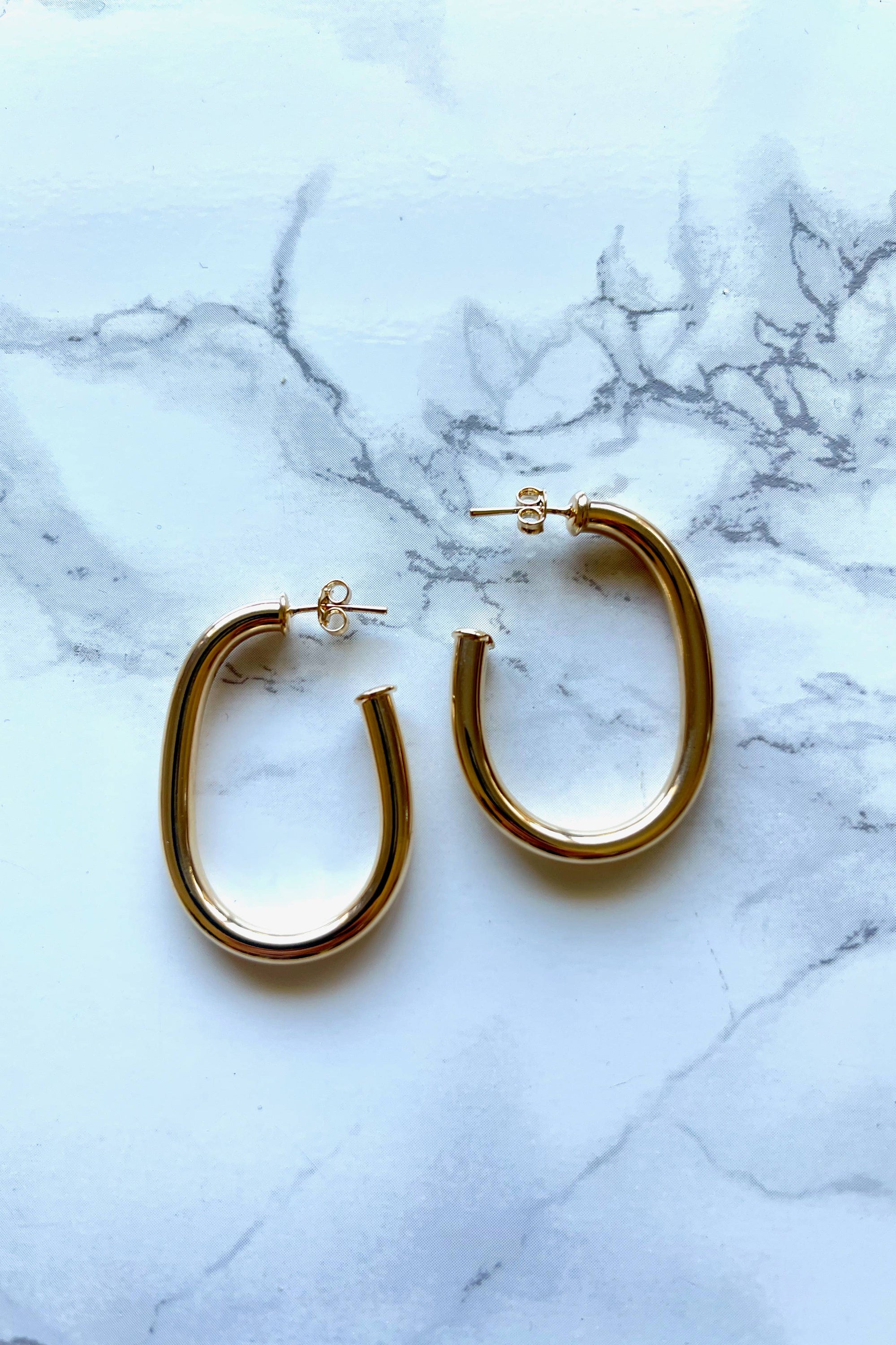 A pair of gold-filled oval thick hoop earrings placed on a marble surface.